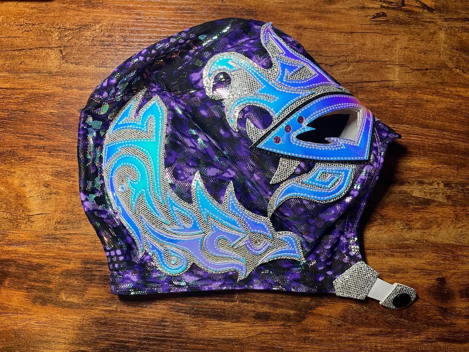 Purple and black lucha libre mask with iridescent blue and silver flame patterns on wood.