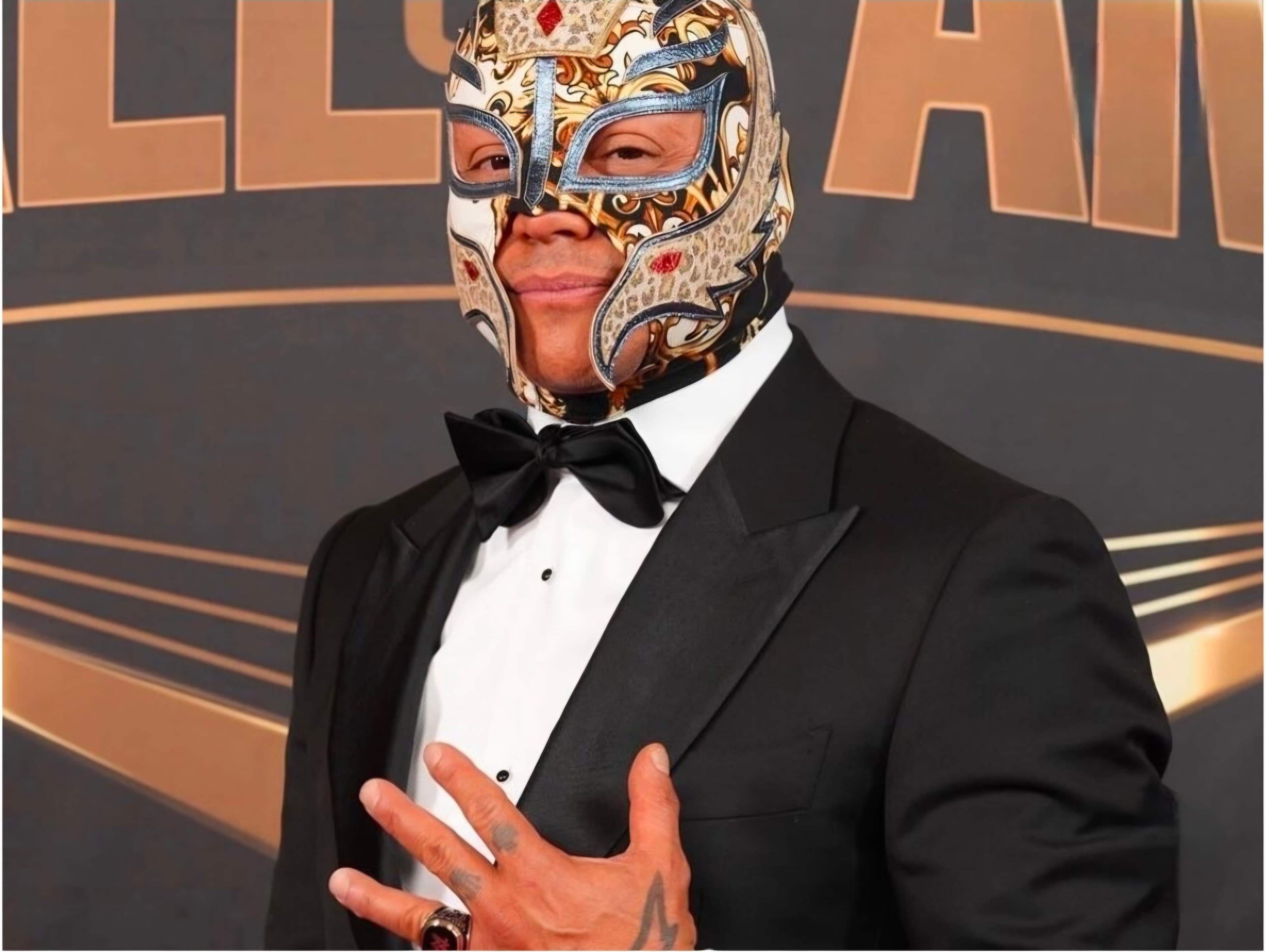 Rey Mysterio in an ornate mask and black tuxedo posing with hand on his chest.