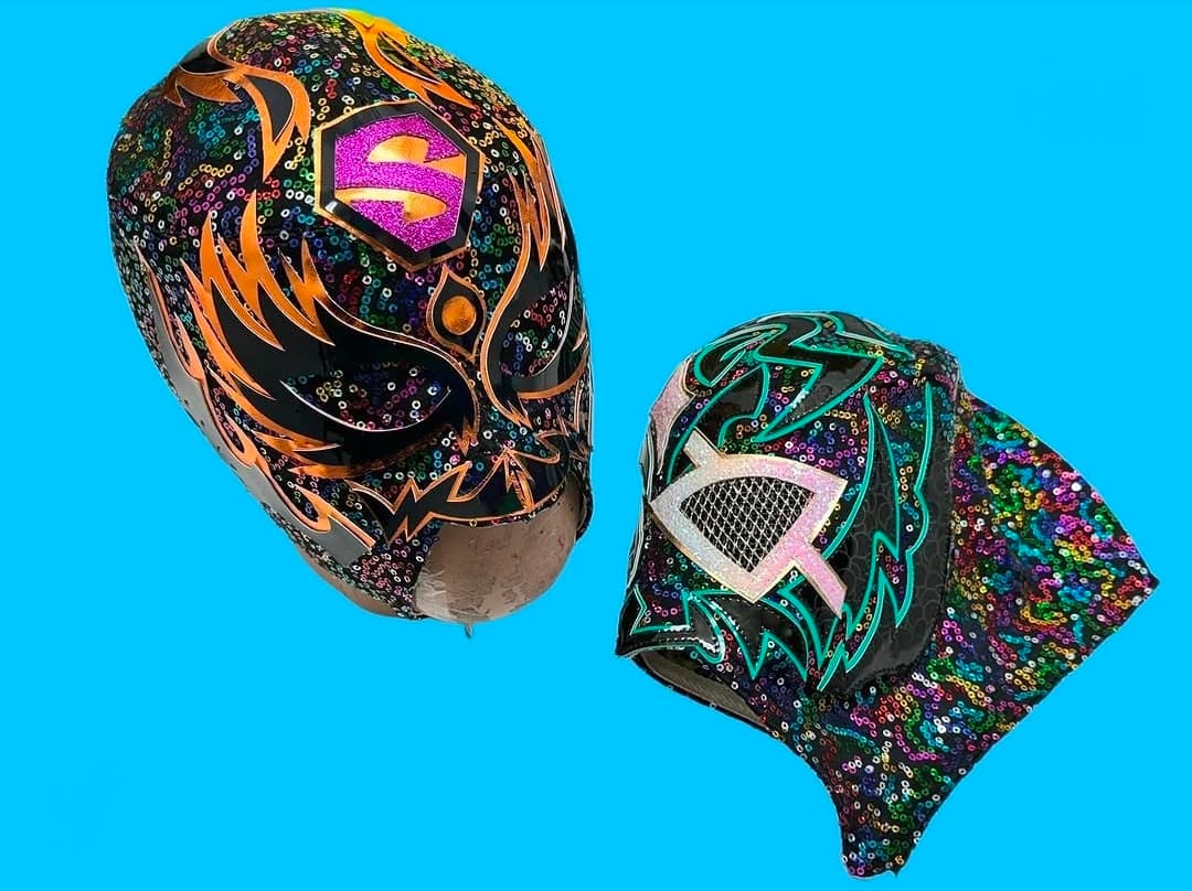 Two vibrant, sequined Lucha Libre masks with intricate patterns against a solid bright blue background.